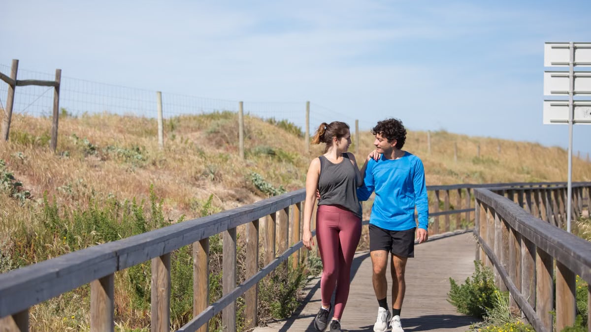A couple in activewear strolling together on a sunny boardwalk in a natural setting.