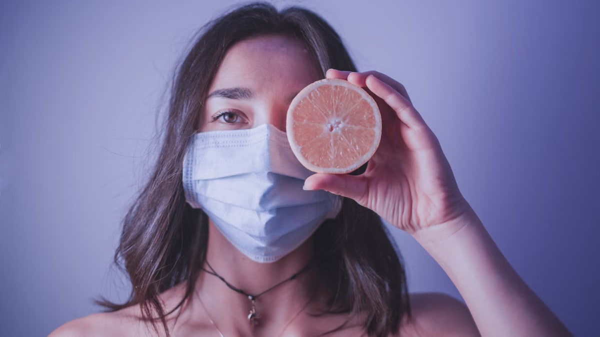 Young female in medical mask looking at camera and covering eye with half of orange while standing near wall during pandemic